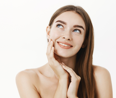 woman-applying-cream-on-face-feeling-skin-soft-and-tender-standing-dreamy-and-delighted-with-result-gazing-at-upper-left-corner-with-sensual-smile-touching-cheek-posing-naked-over-grey-wall